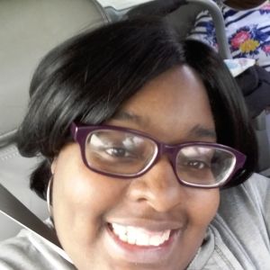 A close up shot of a woman smiling brightly at the camera. She has brown skin and short straight black hair. She is wearing silver hoops and burgundy glasses. She seems happy in the photo.